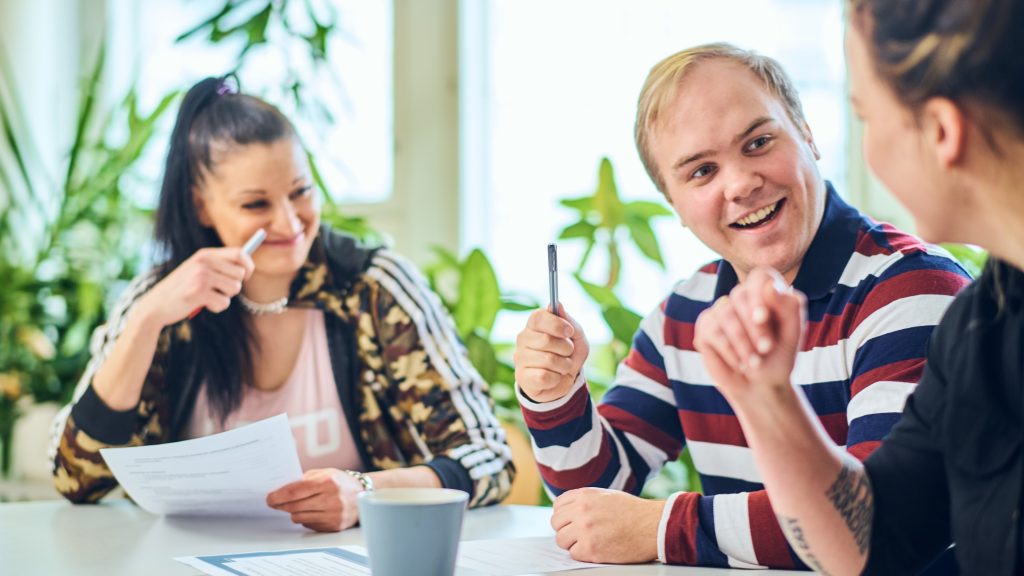 Kolme hymyilevää henkilöä istumassa pöydän äärellä. Pöydällä on papereita ja kyniä.