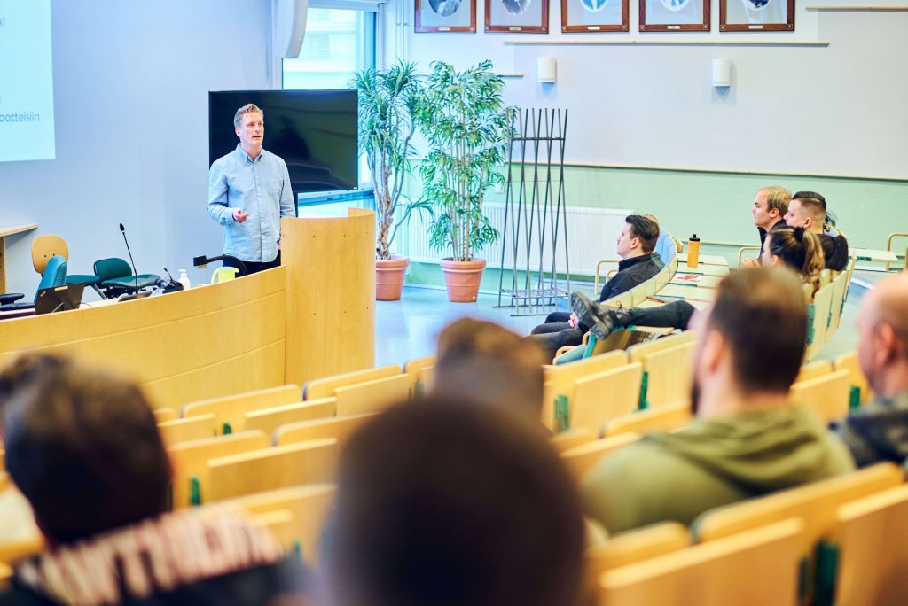 Människor sitter i ett auditorium med många stolar. Framför dem står en person och tittar på de som sitter.
