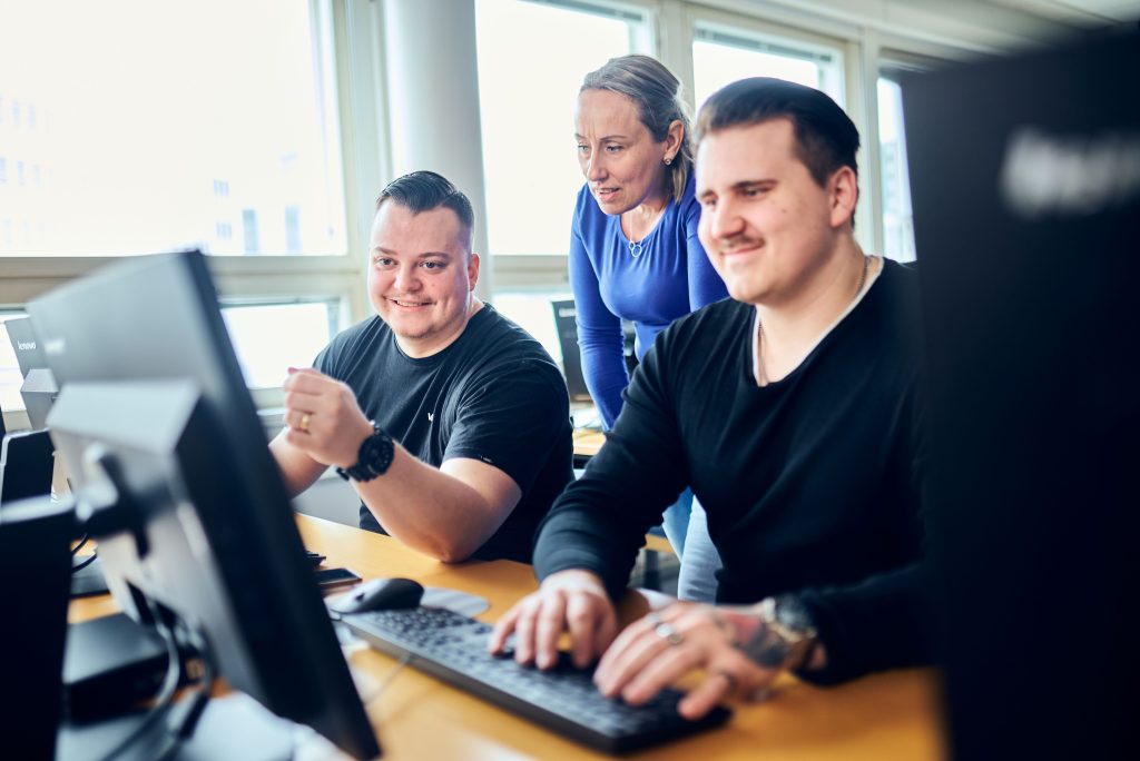 Kaksi hymyilevää henkilöä istumassa tietokoneen äärellä. Yksi seisoo taustalla.