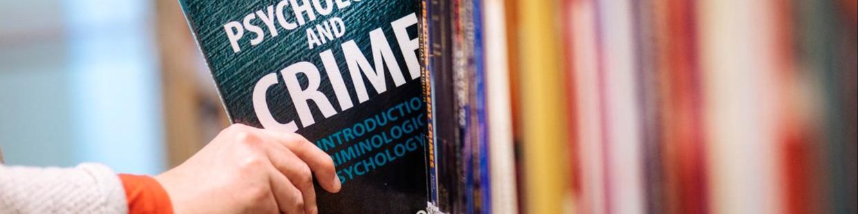 En hand sträcker sig efter en bok med titeln Psychology and Crime som står i bokhylla.