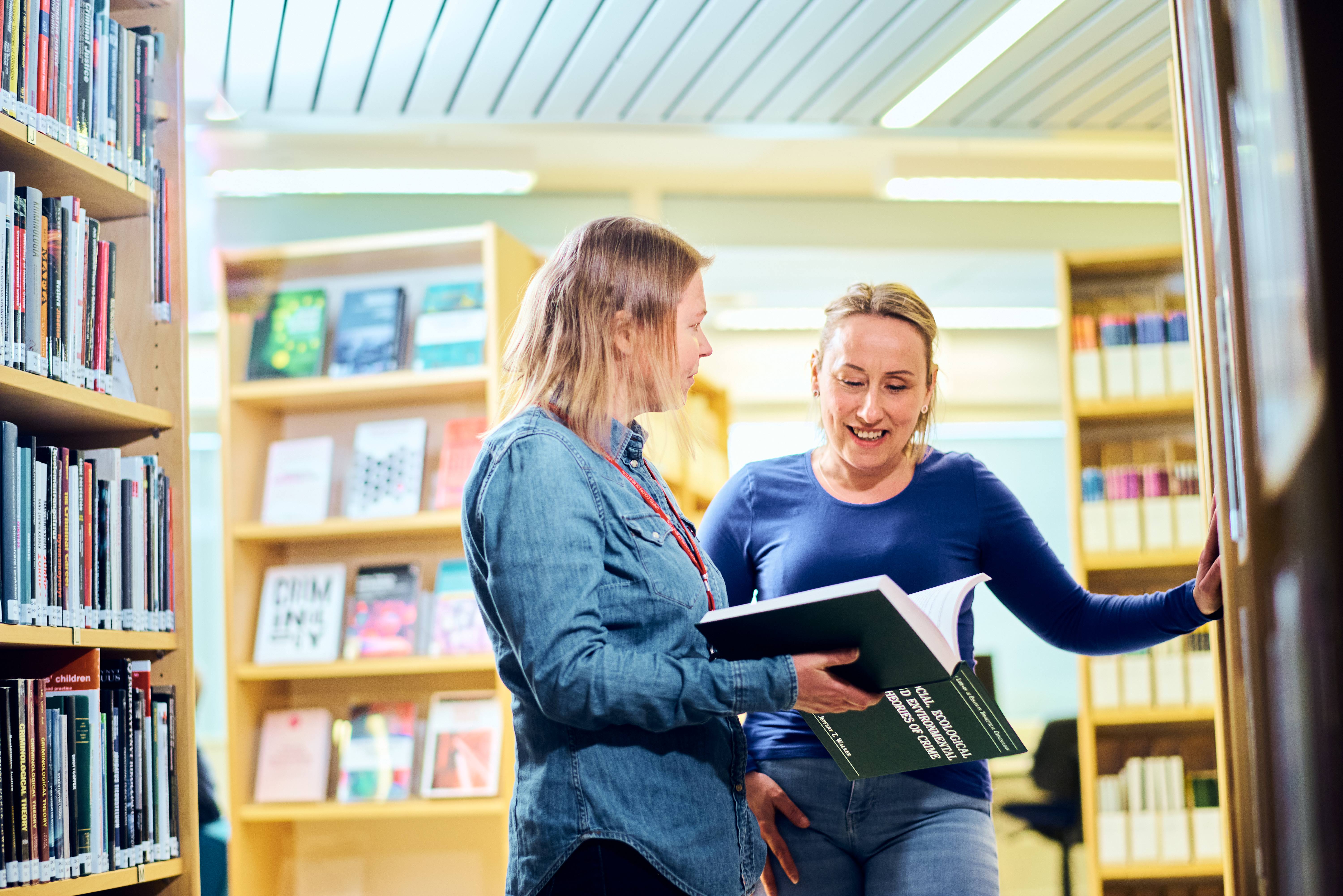 Kaksi naista seisoo kirjahyllyjen edustalla keskustellen kirja käisissään.